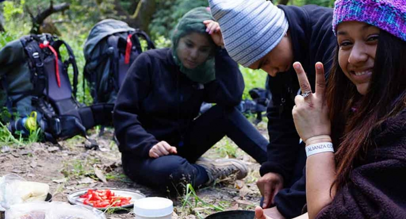 Three outward bound students take a break from backpacking to eat a snack. One of them is giving a peace sign to the camera.